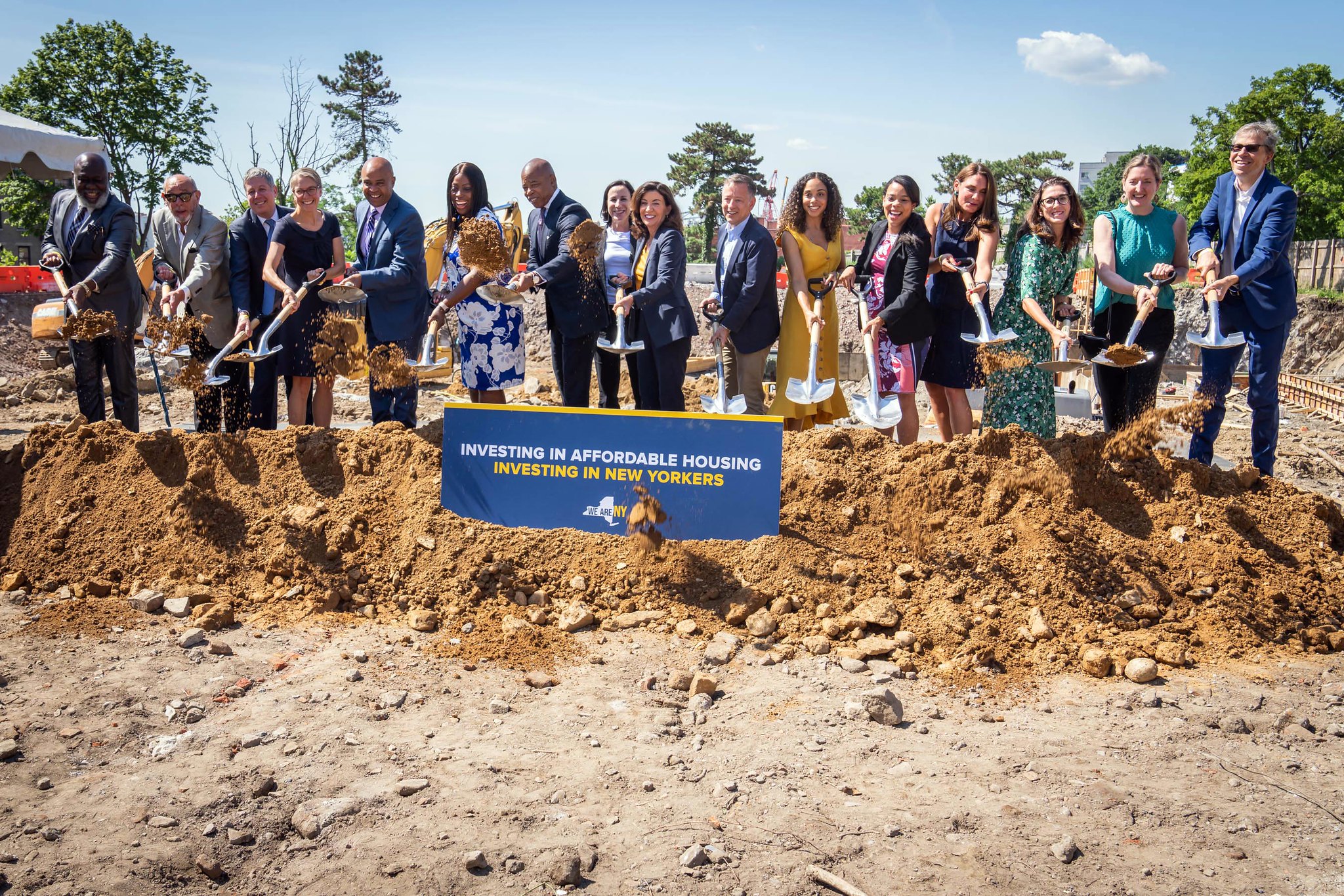 Mayor Eric Adams and Gov. Kathy Hochul, flanked by dozens of people, hold shovels and toss dirt into a pile.