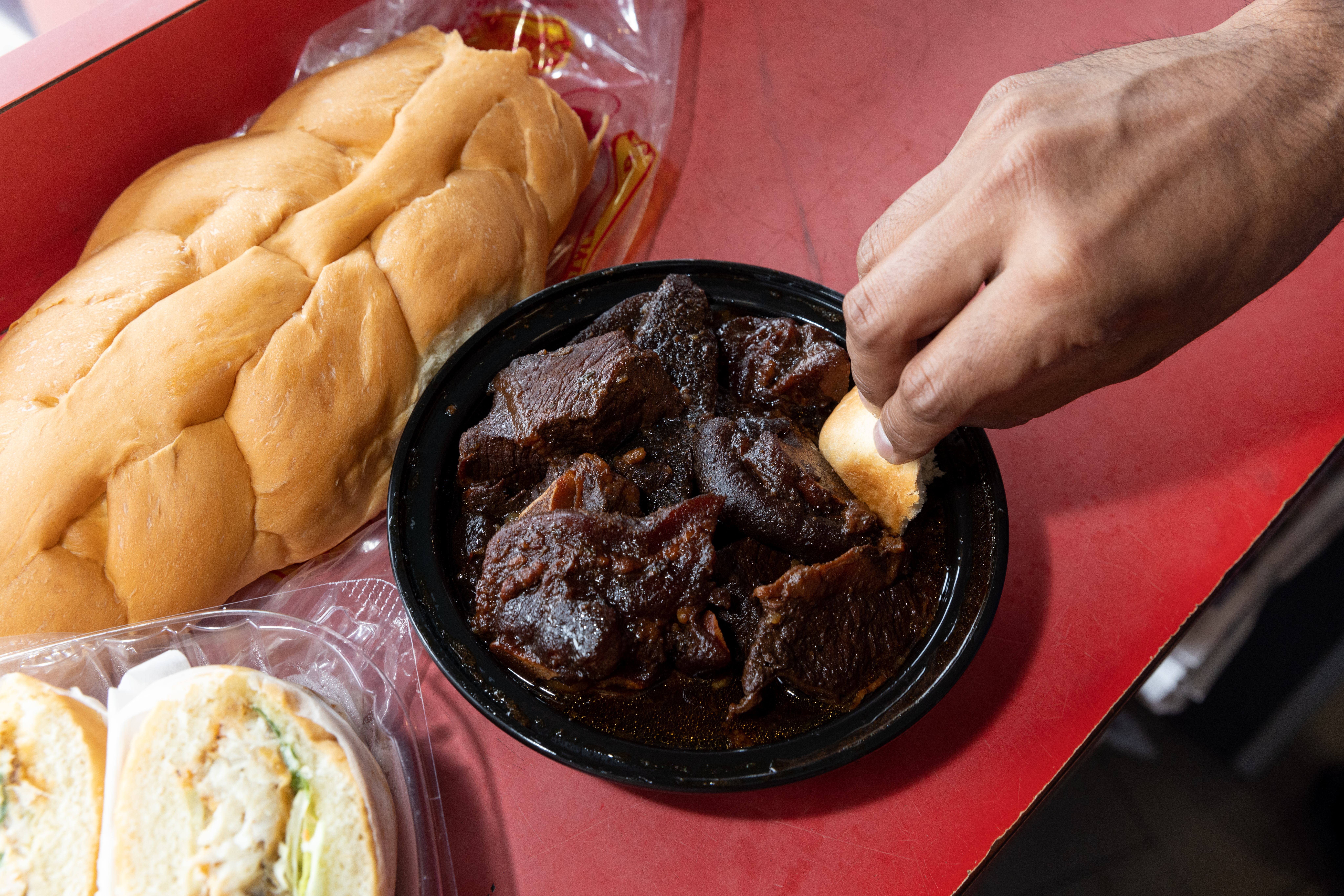 Someone dips Guyana home style bread into a bowl of pepperpot. A loaf of break and a fish sandwich in a container are also on the table.