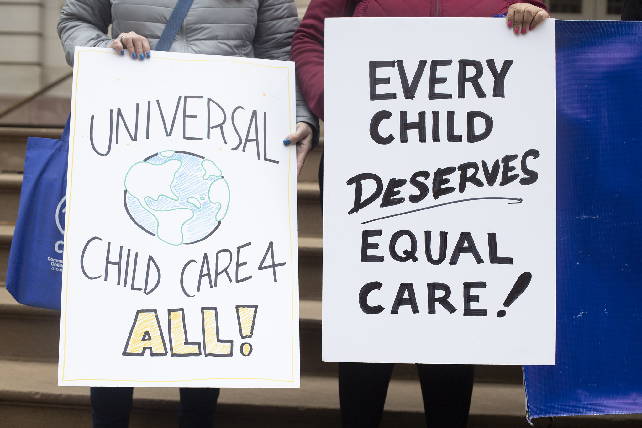 Demonstrators hold signs on the steps of City Hall that say "universal childcare 4 all" and "ever child deserves equal care."