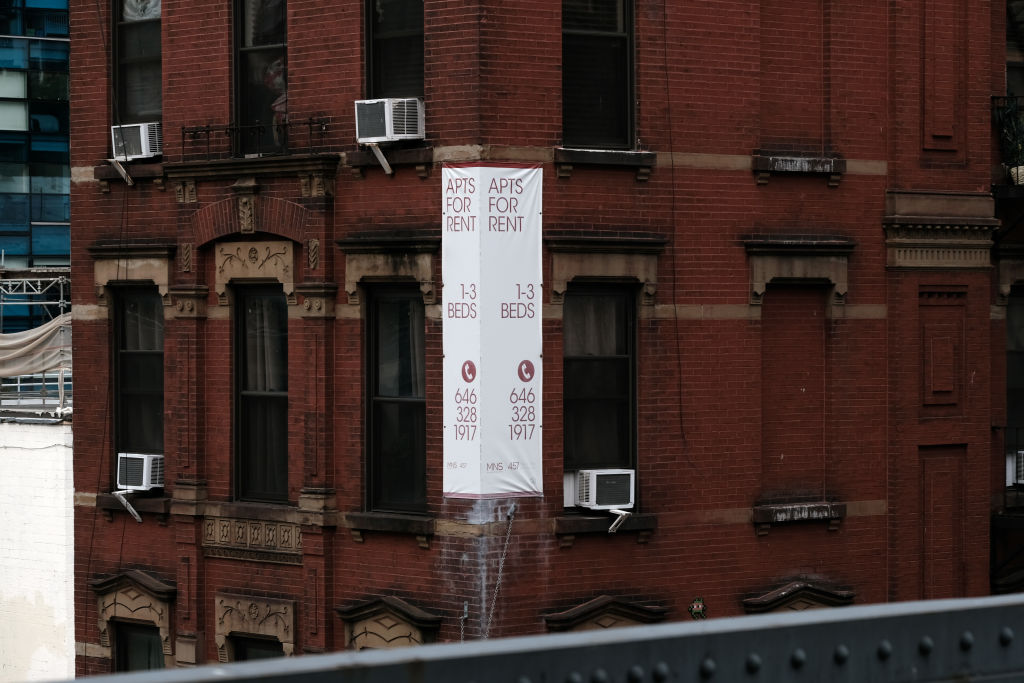 A rent sign advertising apartments hangs on the corner of a brick red building.